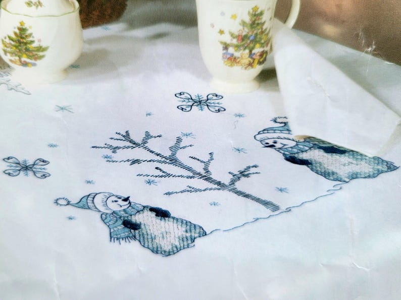 May include: White tablecloth with embroidered winter scene featuring snowmen, snowflakes, and a bare tree in blue and white. A sugar bowl and a mug with a Christmas tree design are in the background.