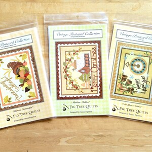 May include: Three vintage postcard quilt patterns featuring Thanksgiving, Christmas, and New Year's themes. Each pattern is printed on a cream-colored card with a brown border and features a colorful illustration. The patterns are from Fig Tree Quilts.