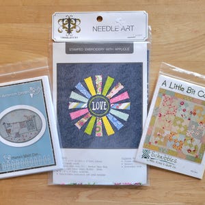 May include: Three craft kits in clear plastic packaging. The center kit is a stamped embroidery with applique, featuring a colorful sunburst design with the word "LOVE". The other two kits are for quilting and embroidery.