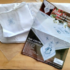 May include: A white tablecloth ensemble with a snowflake design, including a tablecloth, placemat, and napkins. The package includes a tablecloth size of 50 x 70 inches oblong. The package also includes a table runner and a set of napkins. The package is labeled "Blue Snowmen".