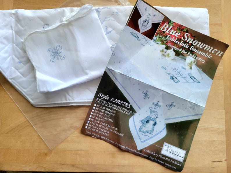 May include: A white tablecloth ensemble with a snowflake design, including a tablecloth, placemat, and napkins. The package includes a tablecloth size of 50 x 70 inches oblong. The package also includes a table runner and a set of napkins. The package is labeled "Blue Snowmen".