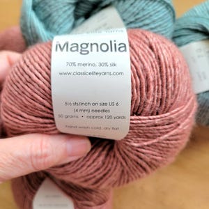 May include: Close-up of several skeins of yarn in shades of pink and teal. The label on the pink yarn reads "Magnolia" and lists the fiber content as 70% merino, 30% silk. The label also indicates 5.5 stitches per inch on US size 6 (4 mm) needles.