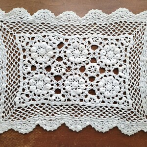 May include: A rectangular white crochet doily featuring a central pattern of six circular floral motifs. The piece is surrounded by a wide, intricate lattice border and finished with a scalloped edge. The design is composed of fine white thread.