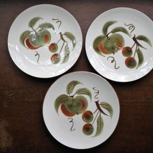 May include: Three white ceramic plates with a hand-painted design of green apples and red cherries. The plates have a brown rim and are arranged on a dark brown surface. The fruit design includes green leaves and brown stems.