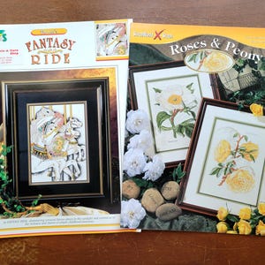 May include: Two cross-stitch pattern books. One features a framed carousel scene with the title "Fantasy Ride." The other book is titled "Roses & Peony" and displays framed floral cross-stitch designs. White flowers and decorative stones surround the books.