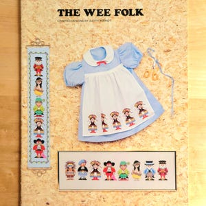 May include: A craft project display featuring a blue and white dress, cross-stitch samplers, and scissors. The main text reads "THE WEE FOLK." The dress has a white apron with embroidered figures. The samplers depict various folk characters.