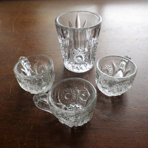 May include: Four clear, vintage glass pieces: a tall tumbler and three small cups with handles. The glassware features a starburst and floral pattern. The objects are arranged on a dark wooden surface.