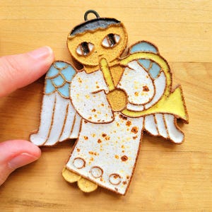 May include: A handcrafted angel ornament with a trumpet, featuring a white robe with brown speckles, blue and white wings, and a yellow trumpet. The angel has a black halo and is held by a hand against a wooden background.