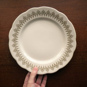 May include: Off-white ceramic plate with a scalloped edge and a decorative border. The border features a repeating pattern in a muted gold color. The plate is round and appears to be clean and unused.