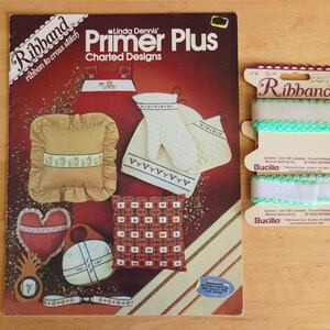 May include: An image featuring a vintage craft pattern book titled "Linda Dennis Primer Plus Charted Designs" with illustrations of cross-stitch projects. Next to it is a spool of white and turquoise Ribband ribbon, perfect for cross-stitch.