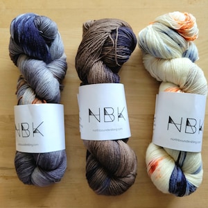 May include: Three skeins of hand-dyed yarn in shades of blue, brown, and cream. Each skein is wrapped with a white label printed with "NBK" and "northboundknitting.com". The yarn is arranged on a light brown wooden surface.