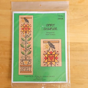 May include: A cross-stitch sampler kit titled "Spirit Sampler" by Diane Arthurs. The design features a star, heart, and bird motif with floral elements. The kit is packaged in a clear plastic sleeve.