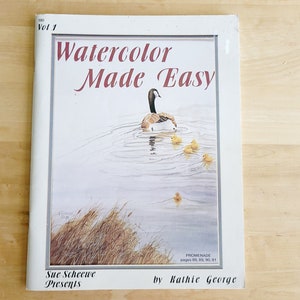 May include: A book titled "Watercolor Made Easy" with a watercolor painting of a goose and ducklings on the cover. The book is white with the title in red and black text. The author's name is Kathie George.