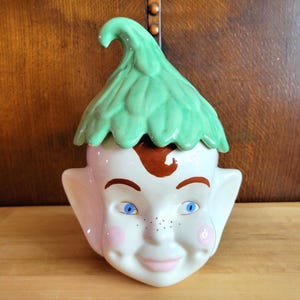 May include: A vintage ceramic cookie jar shaped like a smiling elf face. The jar features a green leafy hat, brown hair, blue eyes, pink cheeks, and freckles. The jar is white with painted details.