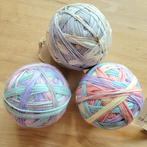 May include: Three colorful yarn balls in pastel shades of blue, pink, purple, and yellow. The yarn is arranged in a crisscross pattern. A small tag with text is attached to one of the balls. The yarn balls are on a wooden surface.