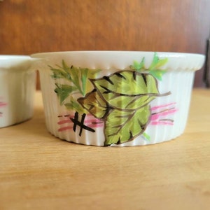 May include: Two white ceramic ramekins with a ribbed exterior. Each ramekin features a hand-painted design of green leaves, brown accents, and pink horizontal lines. The ramekins are set on a light wooden surface.