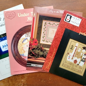 May include: Four rectangular cross stitch pattern booklets featuring various designs. The booklets include titles such as Popcorn Kreations, Under The Mistletoe, Joy to the World, and Heart in Hand with the text In search of the Perfect Snowman.