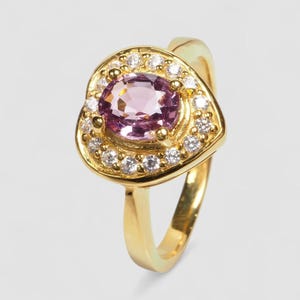 May include: A gold ring featuring a central oval-cut light purple gemstone surrounded by a halo of small, clear, round-cut white stones. The setting is shaped like a rounded triangle, and the band is a smooth, polished gold metal finish.