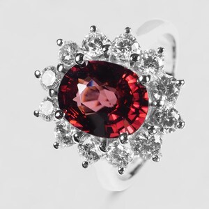 May include: A silver-toned ring featuring a large, oval-cut deep red gemstone at the center. The central stone is surrounded by a halo of twelve small, round, clear faceted stones arranged in a floral pattern, set on a polished metal band.
