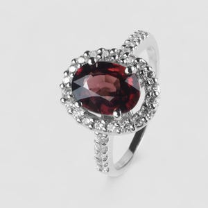 May include: A silver-toned ring featuring a large, oval-cut deep red garnet gemstone at the center. The garnet is surrounded by a halo of small, clear, round-cut stones. The band is also set with a row of small, clear, round-cut stones along the top.