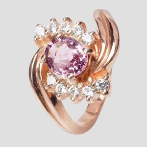 May include: A rose gold ring featuring a central oval-cut pink gemstone surrounded by a halo of small, clear, round-cut white stones. The band has a smooth, polished finish and curves upwards to frame the cluster of stones in a bypass-style setting.