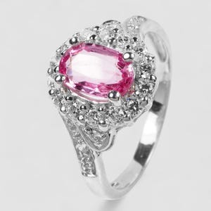 Puede incluir: Un anillo de tono plateado con una piedra preciosa rosa central de corte ovalado, rodeada por un halo de pequeñas piedras claras de corte redondo. La banda también está decorada con pequeñas piedras claras redondas a los lados, creando un brillo.