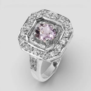 May include: A silver-toned ring featuring a central round, light pink gemstone set within a textured, octagonal frame. The frame is bordered by a halo of small, clear, sparkling stones, with additional clear stones embedded along the ring band.