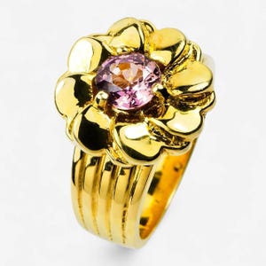 May include: A gold ring featuring a flower-shaped setting with a round, faceted pink gemstone at its center. The band consists of three parallel vertical ridges that extend from the base of the floral design, creating a polished, reflective surface.