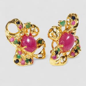 Puede incluir: Un par de pendientes dorados con una gran piedra central rosa ovalada en cabujón rodeada por un marco de metal calado. El marco está decorado con piedras redondas más pequeñas de color rosa, verde y azul oscuro engastadas en la estructura.