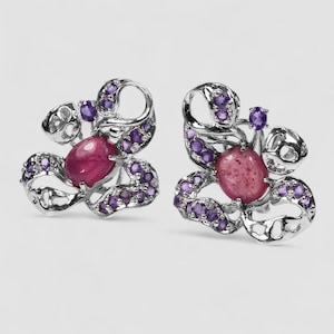 Può includere: Un paio di orecchini color argento con una pietra centrale ovale a cabochon color rubino, circondata da piccole pietre rotonde color ametista incastonate in una montatura in metallo traforato dal design a volute.