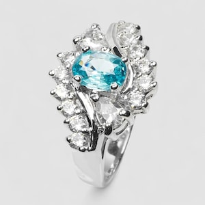 May include: A silver-toned ring featuring a central oval-cut bright blue gemstone surrounded by a cluster of numerous small, clear, round-cut stones. The setting is metallic and polished, with the stones arranged in a dense, layered pattern.