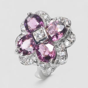 May include: A silver-toned ring featuring a flower-shaped design with four oval-cut purple gemstones arranged around a central clear stone. The petals are accented with small, clear pavé-set stones along the outer edges of the metal setting.