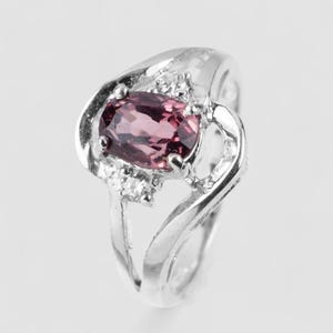 May include: A silver-toned ring featuring a central oval-cut dark pink gemstone held in a prong setting. The band has a split, swirling design and is accented with two small, clear, round-cut stones positioned on either side of the main gem.