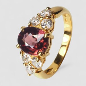 May include: A gold ring featuring a large, oval-cut, deep pinkish-red gemstone at the center, held by four prongs. The band is accented with a cluster of three small, round, clear white stones on each side of the central gemstone setting.