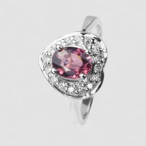 May include: A silver-toned ring featuring a central oval-cut deep pink gemstone. The stone is surrounded by a halo of small, clear, round-cut stones set within a heart-shaped frame. The band is smooth and polished, tapering toward the back of the ring.