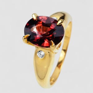 May include: A gold ring featuring a large, oval-cut deep red garnet gemstone held by four prongs. The band is smooth and polished gold, accented with two small, round, clear white diamonds set directly into the metal on either side of the center stone.
