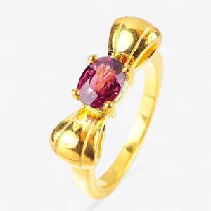 May include: A gold ring featuring a polished yellow gold band and a central bow-shaped setting. The centerpiece is an oval-cut, deep magenta-colored gemstone held in place by four small prongs, creating a distinct contrast against the gold metal.