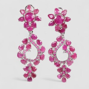 Puede incluir: Un par de pendientes colgantes de tono plateado con múltiples rubíes de color rojo rosado, facetados en forma de pera y redondos, dispuestos en un racimo floral superior y un marco de gota calado, con una sección inferior que cuelga.