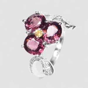 May include: A silver ring featuring three oval-cut deep pink gemstones arranged in a cluster with a small round yellow gemstone at the center. The band is accented with small clear stones along the curved metalwork that extends from the main setting.