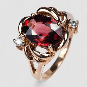 May include: A rose gold ring featuring a large oval-cut deep red garnet gemstone at the center, held by four prongs. The band is designed with intricate, swirling metal loops on each side, accented by a single small, round, clear white diamond.