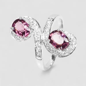 May include: A silver-toned bypass ring featuring two oval-cut deep pink gemstones, each surrounded by a halo of small clear stones. The band is split into an S-shape, with the pavé-set clear stones extending along the curves of the metal frame.