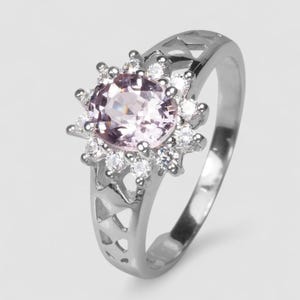 May include: A silver-toned ring featuring a central round, pale pink gemstone surrounded by a halo of small, clear, round-cut stones. The band has a decorative, open-work lattice pattern along the sides, creating a detailed and structured appearance.