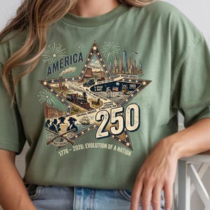 May include: A sage green t-shirt featuring a large star graphic with the text AMERICA and 250. The design includes historical illustrations of a steam train, a ship, a moon landing, and a city skyline, with the text 1776-2026: EVOLUTION OF A NATION.