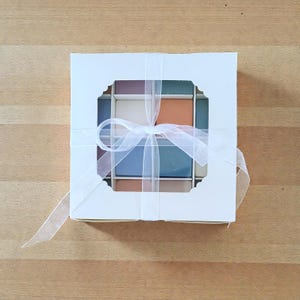 May include: A square white cardboard gift box with a central die-cut window revealing a grid of sixteen small, square, solid-colored items in shades of blue, terracotta, cream, and mauve, tied with a translucent white ribbon in a bow.