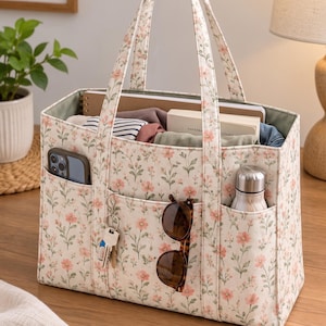 May include: A rectangular fabric tote bag featuring a cream background with a repeating pattern of small pink flowers and green stems. The bag has two top handles, three exterior front pockets, and an open top containing various personal items.