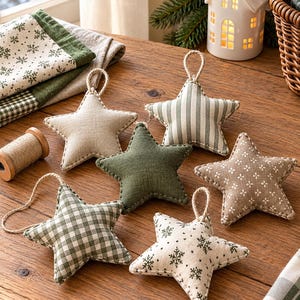 May include: A collection of six fabric star ornaments featuring various patterns including solid olive green, beige, green and white gingham, green stripes, and white with green snowflake prints, each with a jute twine hanging loop.