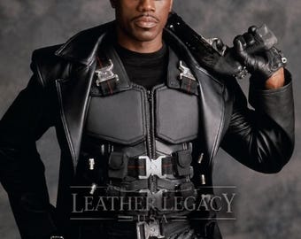 May include: A black leather trench coat worn over a tactical vest with multiple buckles and straps. The outfit includes black gloves and dark sunglasses. The text LEATHER LEGACY USA is visible across the center of the chest area of the garment.