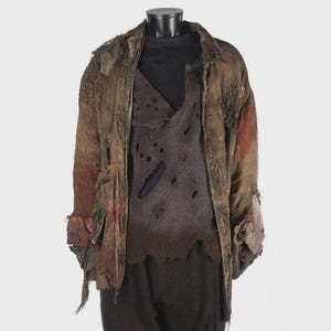 Puede incluir: Una chaqueta marrón desgastada con bordes deshilachados y parches de color marrón rojizo, sobre una camisa gris carbón oscuro con numerosos agujeros pequeños y un dobladillo irregular, junto a pantalones marrón oscuro en un maniquí.