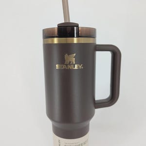 Peut inclure: Un gobelet Stanley Quencher H2.0 marron fonc&eacute; avec un logo dor&eacute;, une poign&eacute;e assortie et un couvercle translucide fonc&eacute; avec une paille. La base pr&eacute;sente une &eacute;tiquette indiquant 40 oz | 1,18 L, pr&eacute;cisant la capacit&eacute; de ce r&eacute;cipient &agrave; boisson.