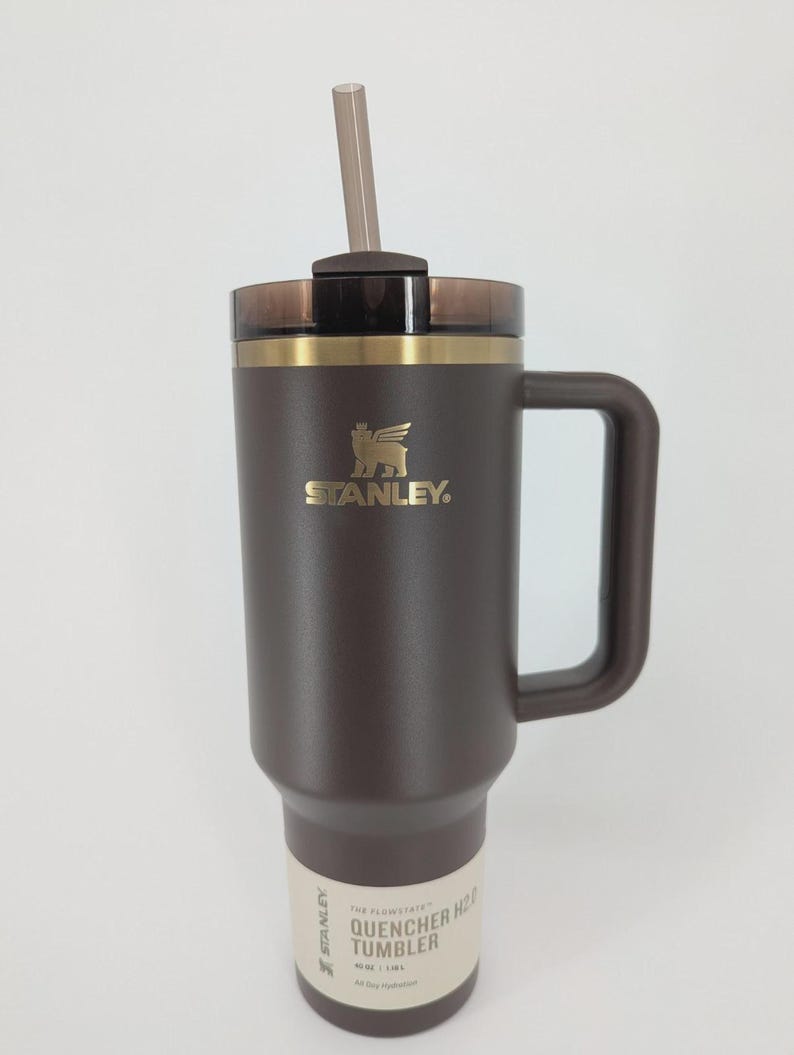 Peut inclure: Un gobelet Stanley Quencher H2.0 marron fonc&eacute; avec un logo dor&eacute;, une poign&eacute;e assortie et un couvercle translucide fonc&eacute; avec une paille. La base pr&eacute;sente une &eacute;tiquette indiquant 40 oz | 1,18 L, pr&eacute;cisant la capacit&eacute; de ce r&eacute;cipient &agrave; boisson.
